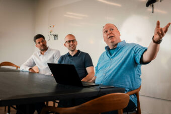 Freek, Sebastiaan en Hans zittend aan zwarte tafel voor een whiteboard kijkend en wijzend naar iets op een beeldscherm buiten de foto