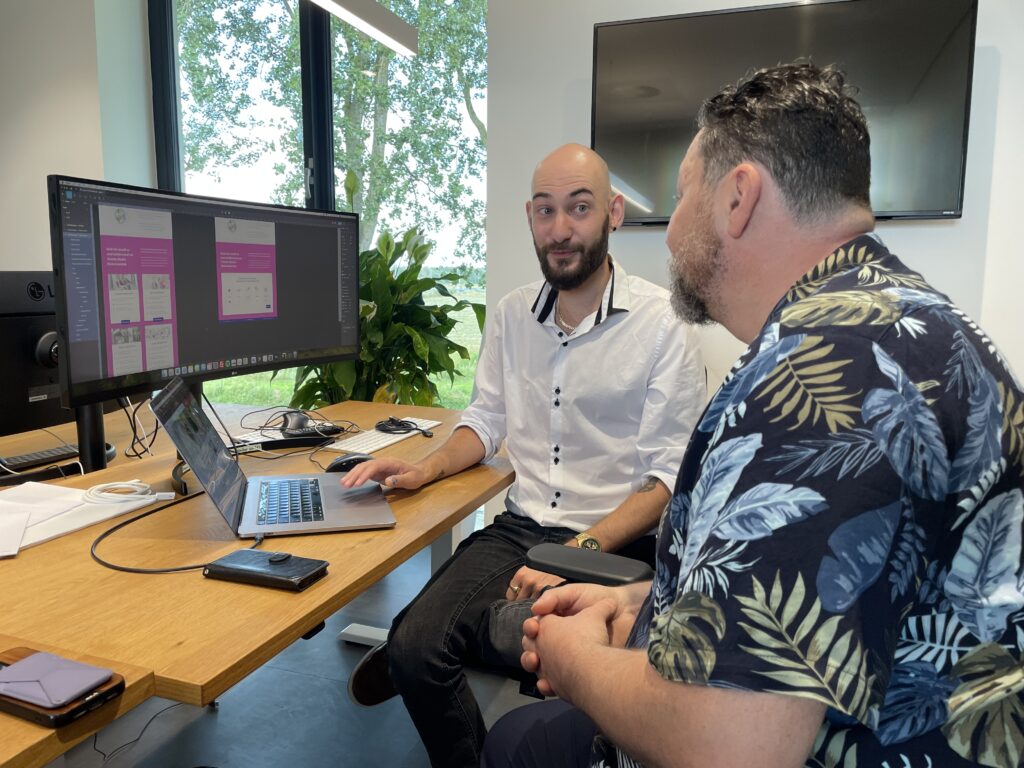 WCAG-Specialist Kelvin Brans in gesprek over een digitaal ontwerp op een computerscherm, met een focus op toegankelijkheid. Op het scherm is een webdesign zichtbaar in paarse tinten, besproken in een kantoor omgeving.