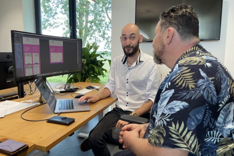 WCAG-Specialist Kelvin Brans in gesprek over een digitaal ontwerp op een computerscherm, met een focus op toegankelijkheid. Op het scherm is een webdesign zichtbaar in paarse tinten, besproken in een kantoor omgeving.