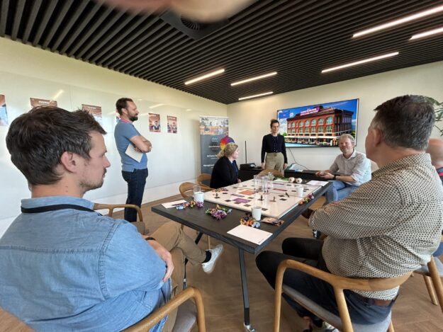 Teamleden spelen Proces Game rond een tafel met speelbord en verschillende attributen en op de achtergrond posters en een beeldscherm