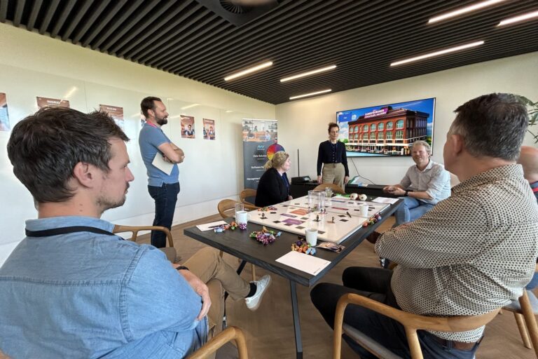 Teamleden spelen Proces Game rond een tafel met speelbord en verschillende attributen en op de achtergrond posters en een beeldscherm